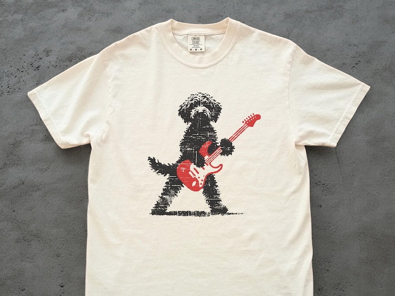 May include: Off-white t-shirt featuring a black dog playing a red electric guitar. The dog is depicted in a stylized, textured design. The guitar has red strings and a white pickguard. The shirt has a crew neck and short sleeves.