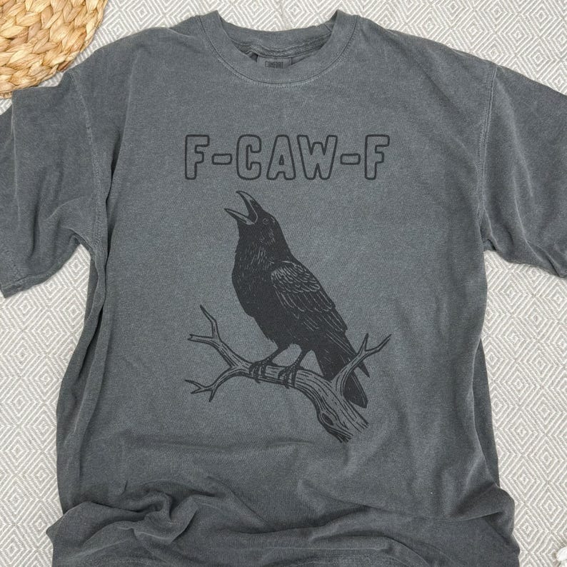 May include: Gray t-shirt with a black graphic of a crow perched on a branch, with the text "F-CAW-F" above the bird. The shirt has a crew neck and short sleeves. The background is a neutral color.