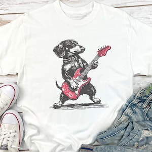 K&ouml;nnte beinhalten: Wei&szlig;es T-Shirt mit einer schwarz-grauen Illustration eines Dackels, der eine rote E-Gitarre spielt. Der Hund tr&auml;gt ein Halsband. Das Shirt wird mit Jeans und wei&szlig;en Turnschuhen gezeigt.