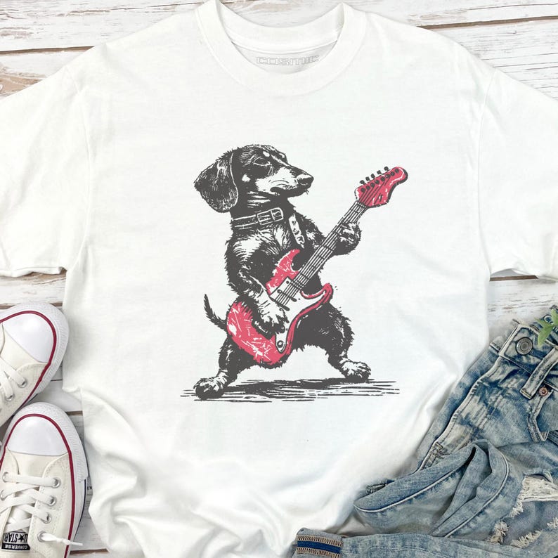 K&ouml;nnte beinhalten: Wei&szlig;es T-Shirt mit einer schwarz-grauen Illustration eines Dackels, der eine rote E-Gitarre spielt. Der Hund tr&auml;gt ein Halsband. Das Shirt wird mit Jeans und wei&szlig;en Turnschuhen gezeigt.