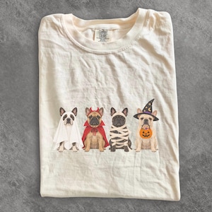 Peut inclure: Un t-shirt couleur crème avec une illustration de quatre bouledogues français en costumes d'Halloween : un fantôme, un diable, une momie et une sorcière tenant un seau citrouille. L'étiquette intérieure du col indique Comfort Colors.