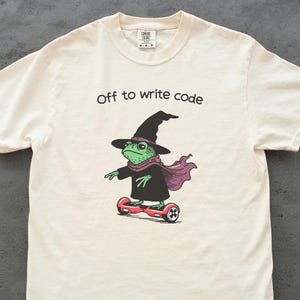 Pode incluir: Camiseta creme com um desenho de um sapo bruxo em uma hoverboard. O sapo verde usa um chapéu e capa pretos, e um cachecol roxo. O texto "Off to write code" está acima. Design divertido.