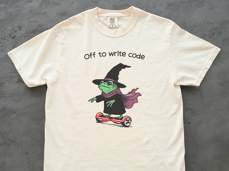 以下が含まれることがあります： オフホワイトのTシャツ。ホバーボードに乗ったカエルの魔女の漫画が描かれています。カエルは緑色で、黒い帽子とマント、紫色のスカーフを着用しています。「Off to write code」の文字が上にあります。面白いデザインです。