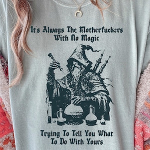 May include: A light blue t-shirt with a dark green illustration of a wizard holding a vial and a bagpipe. The text on the shirt reads "It's Always The Motherfuckers With No Magic Trying To Tell You What To Do With Yours."