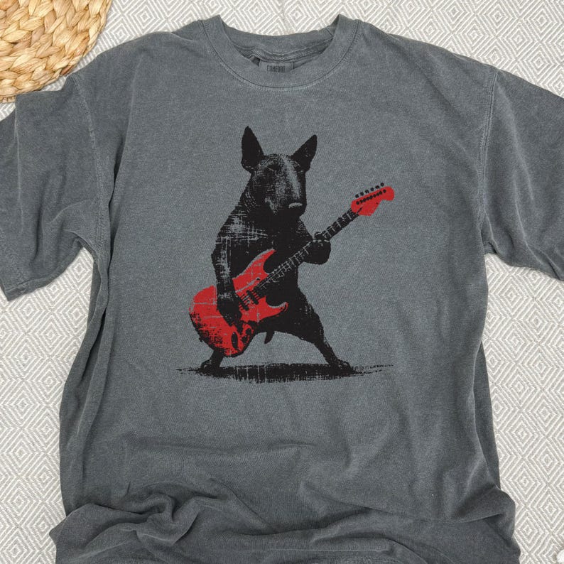 K&ouml;nnte beinhalten: Graues T-Shirt mit einer schwarzen Illustration eines Bullterriers, der eine rote E-Gitarre spielt. Das Design hat einen Used-Look. Das Shirt besteht aus weichem Material und hat einen Rundhalsausschnitt. Ein l&auml;ssiges, musikalisches Kleidungsst&uuml;ck.