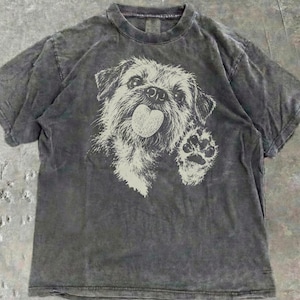 May include: A charcoal gray short-sleeve t-shirt with a washed-out, mottled texture. The center features a cream-colored graphic illustration of a scruffy dog with its tongue hanging out, raising one paw in a waving gesture.