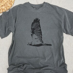 May include: A gray t-shirt featuring a black graphic of a hawk in flight. The bird's wings are spread wide, showcasing detailed feather patterns. The shirt has a crew neck and short sleeves, made of a soft, comfortable material.