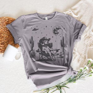 Pu&ograve; includere: Una maglietta grigia con un'illustrazione in bianco e nero di un cane che indossa un cappello da cowboy e siede su una sedia da campeggio sotto una luna piena e stelle. Il cane &egrave; circondato da cactus e un UFO.