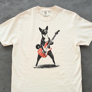 May include: Cream-colored t-shirt featuring a black and white dog playing a red electric guitar. The dog is standing with its legs spread, and the guitar is in front of it. The shirt has a classic crew neck.