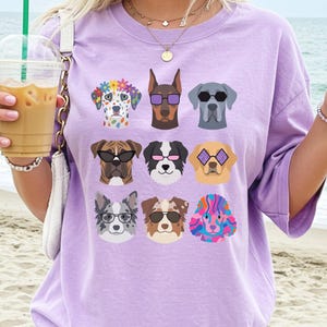 May include: A light purple t-shirt featuring a graphic print of nine different dog breeds arranged in a grid. Each dog is depicted with unique accessories, including colorful sunglasses, a flower crown, and vibrant, multi-colored fur patterns.