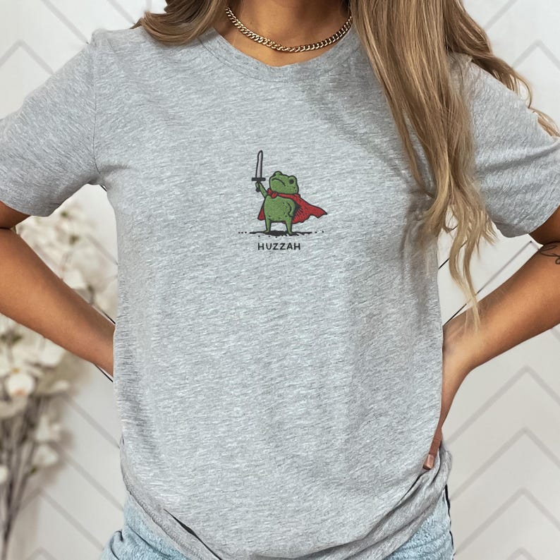May include: A light gray t-shirt featuring a cartoon frog warrior with a sword and red cape. The frog is holding a sword and the word "Huzzah" is printed below the frog.