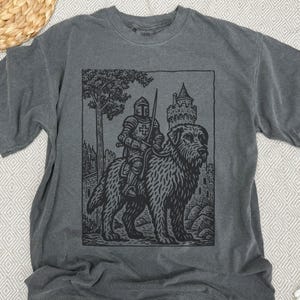 May include: Dark gray t-shirt featuring a black graphic of a knight riding a large dog, with a castle and tree in the background. The knight is wearing armor and holding a sword. The design has a vintage, woodcut style.