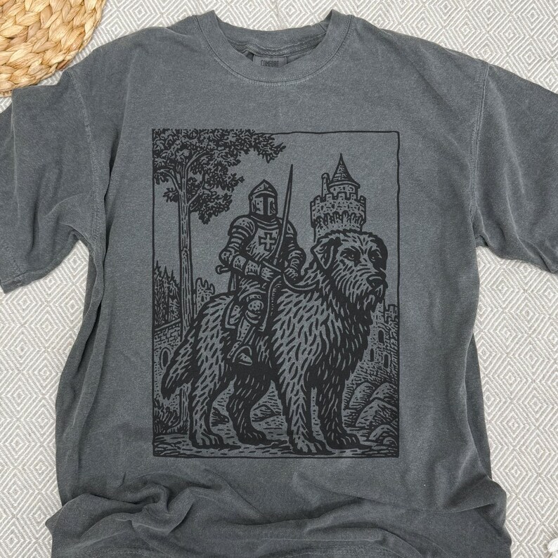 May include: Dark gray t-shirt featuring a black graphic of a knight riding a large dog, with a castle and tree in the background. The knight is wearing armor and holding a sword. The design has a vintage, woodcut style.