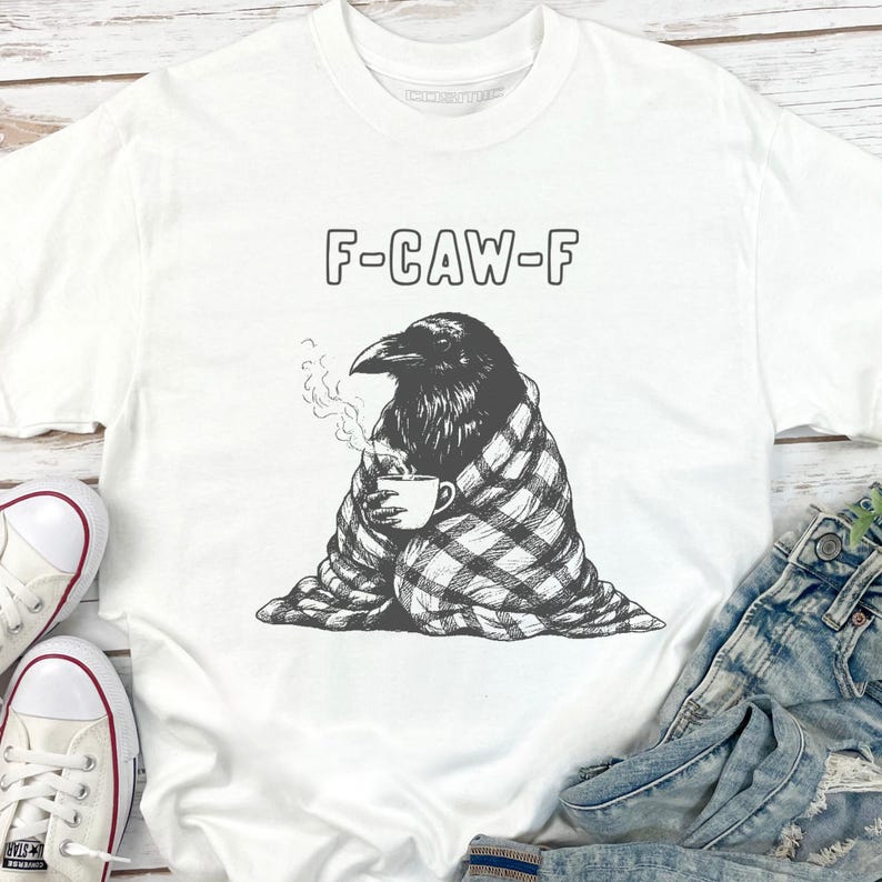 May include: White t-shirt with a graphic of a raven wrapped in a blanket, holding a coffee cup. The text "F-CAW-F" is above the raven. The raven is black and white, and the blanket has a plaid pattern. The shirt is paired with blue jeans and white sneakers.