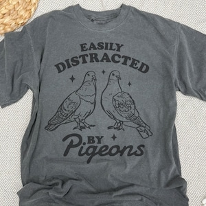 May include: Charcoal grey t-shirt with the phrase "EASILY DISTRACTED BY PIGEONS" in black. The design includes two pigeon illustrations and star shapes. The shirt has a crew neck and short sleeves.