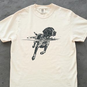 Puede incluir: Camiseta color crema con una ilustración en blanco y negro de un perro nadando en el agua. El perro, de raza pointer, tiene un pelaje manchado y una expresión alerta. El gráfico está centrado en la camiseta.