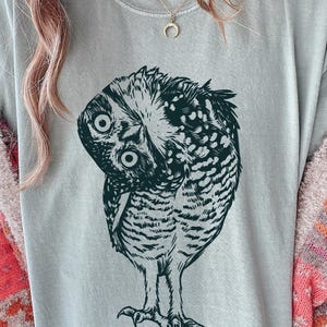 Może przedstawiać: Jasnoszara koszulka z szczeg&oacute;łową ilustracją sowy. Sowa jest przedstawiona w odcieniach ciemnej zieleni i bieli, ze skomplikowanymi wzorami pi&oacute;r. Koszulka jest w stylu casual.