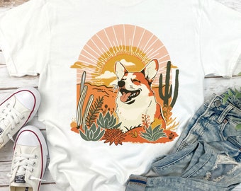 Pode incluir: Camiseta branca com um gráfico de um cão corgi sorridente em uma paisagem desértica com cactos e um pôr do sol. O corgi está rodeado de flores e plantas.