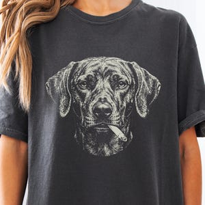 May include: A charcoal grey short-sleeved t-shirt featuring a detailed, monochromatic line-art illustration of a dog's face centred on the chest. The dog is depicted with a cigarette held in its mouth, rendered in a stark white graphic style.
