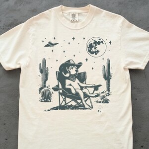 Pu&ograve; includere: Una t-shirt color crema con un'illustrazione in bianco e nero di un cane che indossa un cappello da cowboy e siede su una sedia da campeggio. Il cane &egrave; circondato da cactus e stelle. La luna &egrave; visibile sullo sfondo.