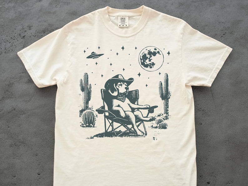 Pu&ograve; includere: Una t-shirt color crema con un'illustrazione in bianco e nero di un cane che indossa un cappello da cowboy e siede su una sedia da campeggio. Il cane &egrave; circondato da cactus e stelle. La luna &egrave; visibile sullo sfondo.
