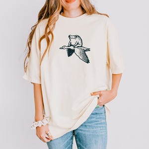 K&ouml;nnte beinhalten: Ein cremefarbenes Kurzarm-T-Shirt mit einem schwarzen Grafikdruck, der einen Frosch auf dem R&uuml;cken einer fliegenden Gans zeigt. Das Shirt hat einen Rundhalsausschnitt und eine lockere Passform mit einer einfachen Strichzeichnung im Brustbereich.