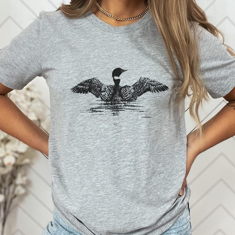 May include: A heather gray t-shirt featuring a black and white graphic of a loon with outstretched wings. The bird is depicted in water, with detailed feather patterns. The shirt has a crew neck and short sleeves.