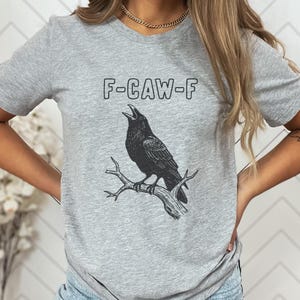 May include: A gray t-shirt featuring a black illustration of a crow perched on a branch, with the text "F-CAW-F" above the bird. The design is printed in black ink.