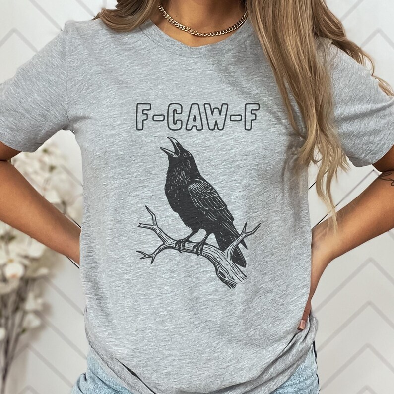 May include: A gray t-shirt featuring a black illustration of a crow perched on a branch, with the text "F-CAW-F" above the bird. The design is printed in black ink.