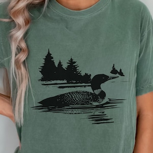 May include: Sage green t-shirt with a black illustration of a loon on a lake, with pine trees in the background. The shirt has a relaxed fit and short sleeves. The design is a nature-themed illustration.