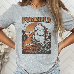 May include: Grey t-shirt with a graphic of a Pomeranian dog as Godzilla, with the text "POMZILLA" in orange. The dog is breathing fire onto a car and buildings, with a sunset in the background.