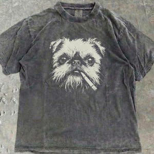 Może przedstawiać: Grafitowy t-shirt z białym nadrukiem przedstawiającym psa z papierosem w ustach. Koszulka ma okrągły dekolt i krótkie rękawy. Tkanina ma wyblakły, vintage wygląd.