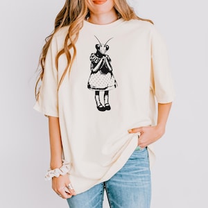 Puede incluir: Camiseta color crema con una ilustraci&oacute;n negra de una mantis religiosa con un vestido. El insecto est&aacute; representado con una cabeza detallada, antenas y un vestido de lunares. La camiseta tiene mangas cortas y un corte holgado, adecuado para uso casual.