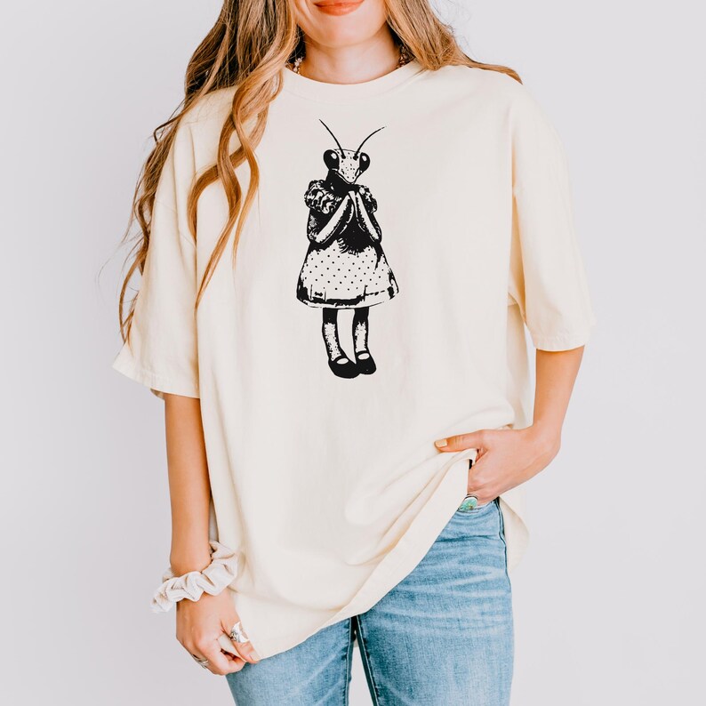 Puede incluir: Camiseta color crema con una ilustraci&oacute;n negra de una mantis religiosa con un vestido. El insecto est&aacute; representado con una cabeza detallada, antenas y un vestido de lunares. La camiseta tiene mangas cortas y un corte holgado, adecuado para uso casual.