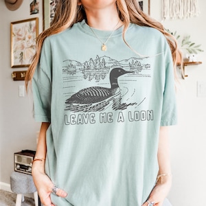 Puede incluir: Camiseta verde claro con una ilustraci&oacute;n en blanco y negro de un somorgujo nadando en un lago, con &aacute;rboles y monta&ntilde;as al fondo. El texto "LEAVE ME A LOON" est&aacute; impreso debajo.