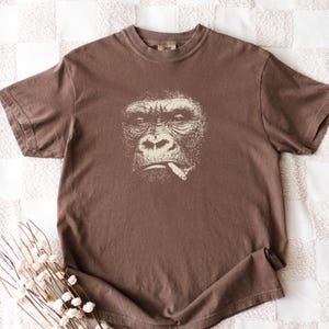 May include: Brown t-shirt featuring a detailed graphic of a gorilla smoking a cigarette. The gorilla's face is rendered in a detailed, off-white illustration. The shirt has a classic crew neck and short sleeves, made of a soft, comfortable material.