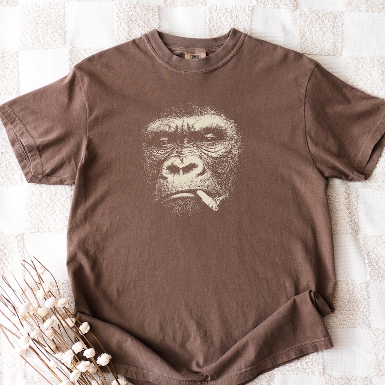 May include: Brown t-shirt featuring a detailed graphic of a gorilla smoking a cigarette. The gorilla's face is rendered in a detailed, off-white illustration. The shirt has a classic crew neck and short sleeves, made of a soft, comfortable material.