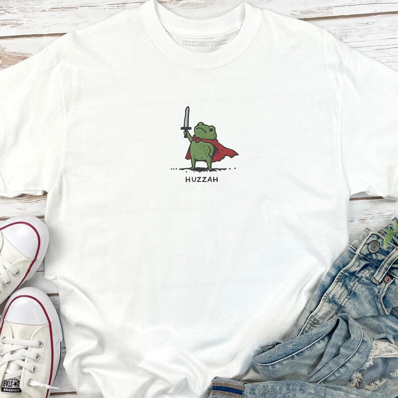 May include: White t-shirt featuring a cartoon frog wearing a red cape and holding a sword. The frog is green with black eyes. The word "Huzzah" is printed below the frog in black letters. The shirt is paired with blue jeans and white sneakers.