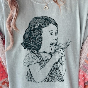 May include: A light blue t-shirt features a black and white illustration of a girl with her mouth open, holding a slug. The girl's dress has a floral pattern. The shirt has a crew neck and short sleeves.