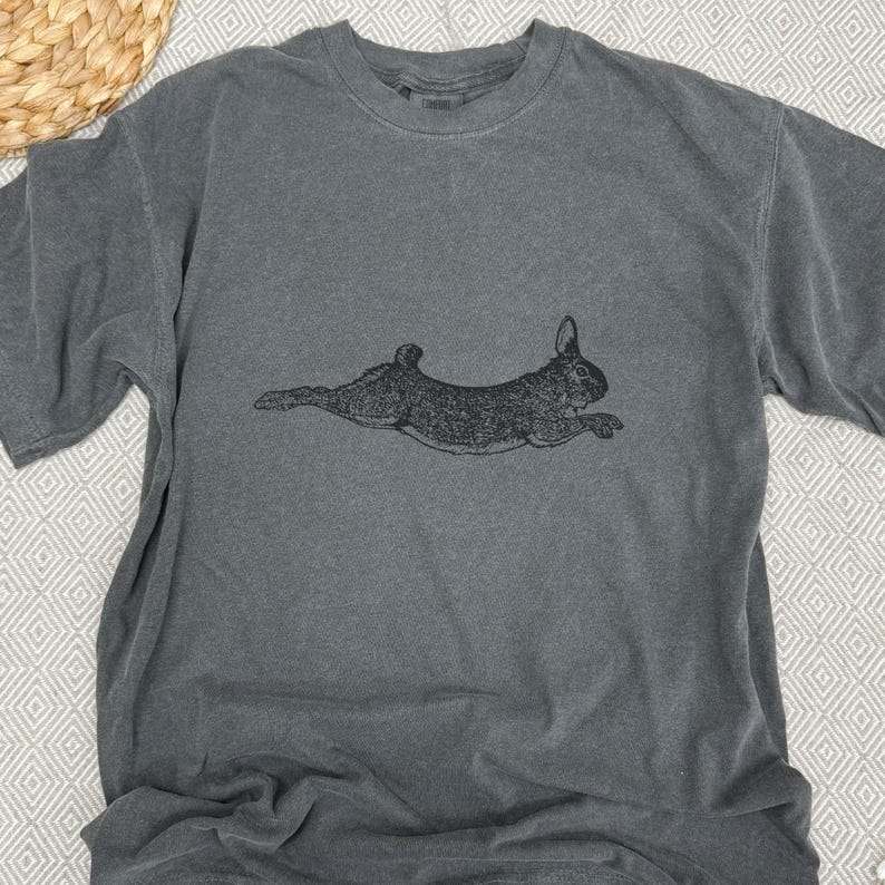 Puede incluir: Una camiseta gris con una ilustraci&oacute;n negra de un conejo corriendo. La camiseta de manga corta tiene cuello redondo y un corte relajado. El dise&ntilde;o del conejo est&aacute; centrado en la parte delantera de la camiseta.