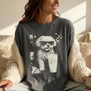 May include: Dark grey t-shirt with a white graphic of a dog wearing sunglasses and drinking from a cup with a straw. The dog is sitting. The shirt has a relaxed fit.