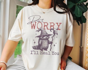 May include: A white t-shirt with a graphic of a raccoon wearing a witch's hat and holding a staff. The text "Don't Worry I'll Heal You" is printed below the graphic.