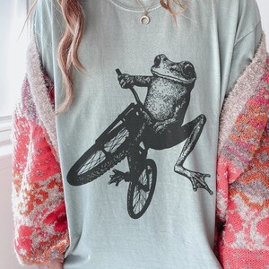 Pu&ograve; includere: Una maglietta azzurra con una grafica nera di una rana in bicicletta. La rana &egrave; seduta, con le gambe distese. La bicicletta &egrave; anch'essa nera. La maglietta ha un girocollo e maniche corte.