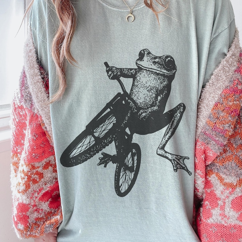 Pu&ograve; includere: Una maglietta azzurra con una grafica nera di una rana in bicicletta. La rana &egrave; seduta, con le gambe distese. La bicicletta &egrave; anch'essa nera. La maglietta ha un girocollo e maniche corte.