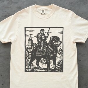Könnte beinhalten: Cremefarbenes T-Shirt mit einem Schwarz-Weiß-Grafik eines Ritters in Rüstung, der auf einem großen Hund, möglicherweise einem Rottweiler, reitet. Der Ritter hält einen Speer, und ein Gebäude ist im Hintergrund. Das T-Shirt hat einen Rundhalsausschnitt und kurze Ärmel.