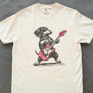 K&ouml;nnte beinhalten: Cremefarbenes T-Shirt mit einer schwarz-wei&szlig;en Illustration eines Dackels, der eine rote E-Gitarre spielt. Der Hund tr&auml;gt ein Halsband. Das Shirt hat einen Rundhalsausschnitt und kurze &Auml;rmel.