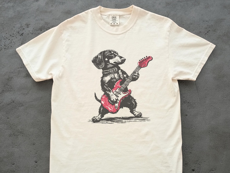 K&ouml;nnte beinhalten: Cremefarbenes T-Shirt mit einer schwarz-wei&szlig;en Illustration eines Dackels, der eine rote E-Gitarre spielt. Der Hund tr&auml;gt ein Halsband. Das Shirt hat einen Rundhalsausschnitt und kurze &Auml;rmel.
