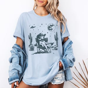 Pu&ograve; includere: Una maglietta blu chiaro con una grafica di un cane che indossa un cappello da cowboy e siede su una sedia da campeggio sotto una luna piena e stelle. Il cane &egrave; circondato da cactus e un UFO vola sopra.