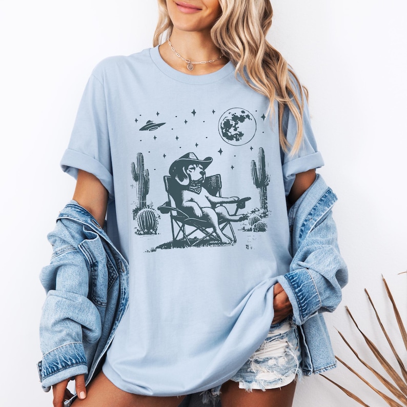 Pu&ograve; includere: Una maglietta blu chiaro con una grafica di un cane che indossa un cappello da cowboy e siede su una sedia da campeggio sotto una luna piena e stelle. Il cane &egrave; circondato da cactus e un UFO vola sopra.