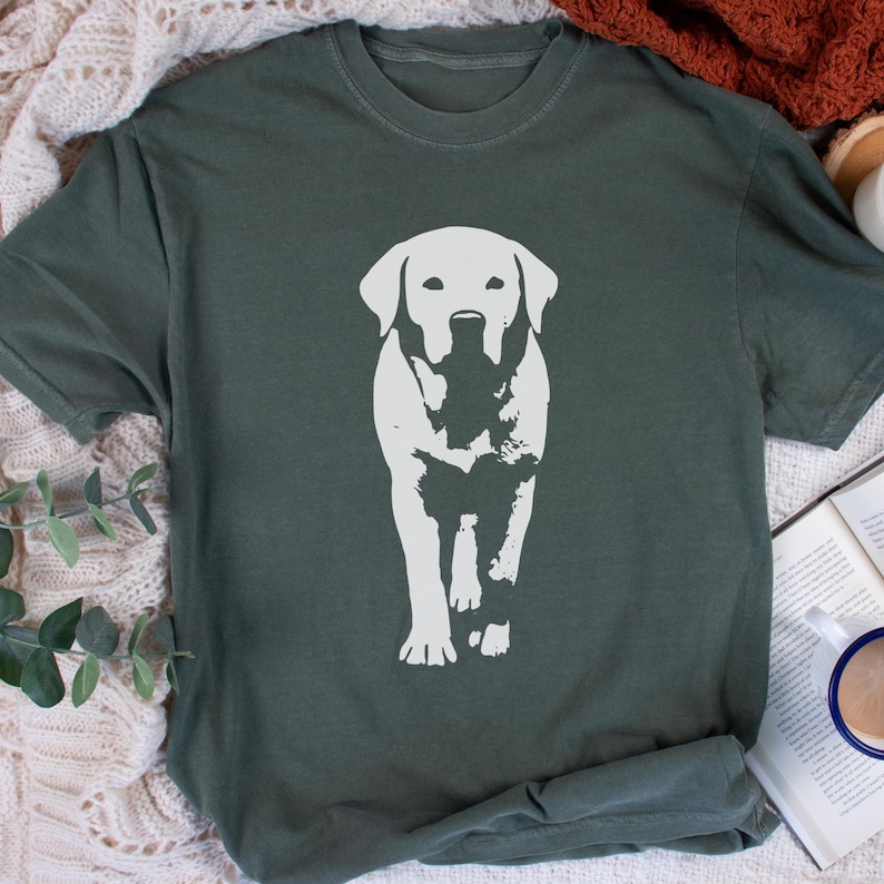 May include: A dark green t-shirt featuring a white graphic of a Labrador Retriever dog. The dog is depicted in a standing pose, with a detailed outline. The shirt has a crew neck and short sleeves, and is made of a soft material.